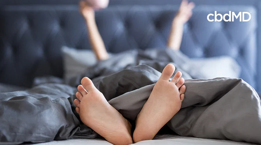 Bare feet in bed with gray bedding, relaxed morning, sleep wellness concept, cbdMD logo