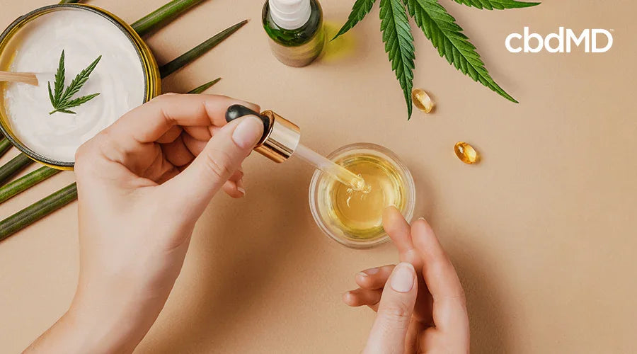 Hands using CBD oil dropper with hemp leaves, capsules, and lotion on beige background