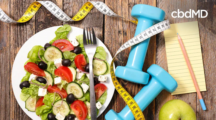 Fresh vegetable salad, blue dumbbells, measuring tape, notepad, and apple on wooden table for healthy lifestyle