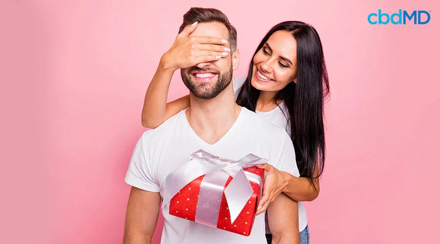 A woman behind a man covering his eyes with one hand and holding a gift in front of him in her other hand - cbdMD