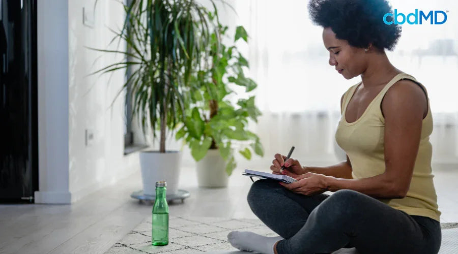 Woman sitting on floor writing healthy new year resolutions, water bottle and plant nearby