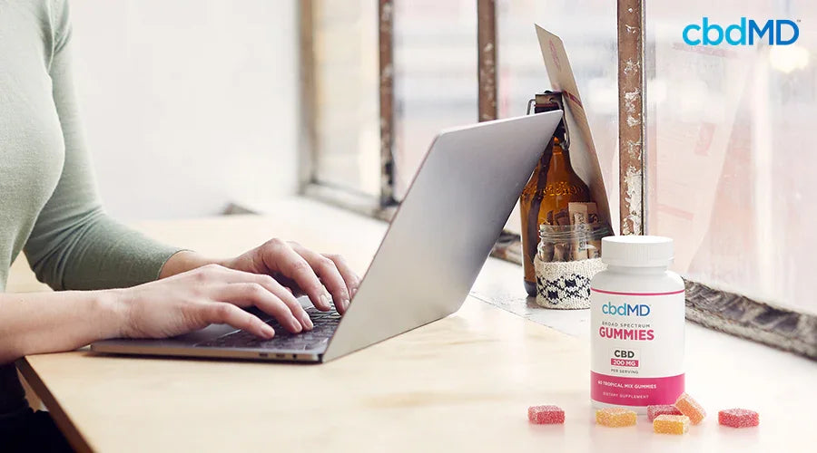Person working on laptop at desk with CBD gummies bottle and assorted gummies nearby