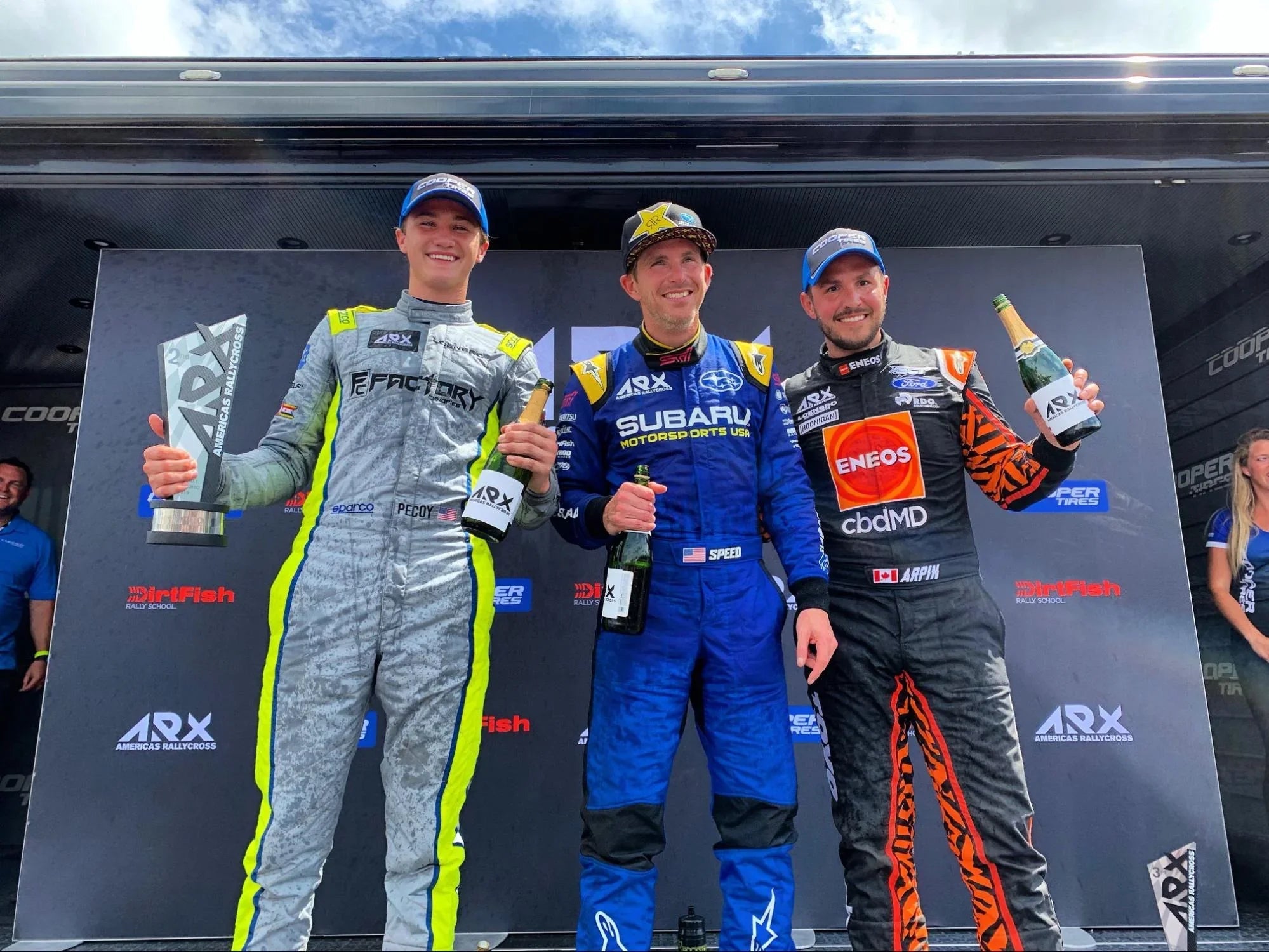 Three rallycross drivers in racing suits celebrating on the podium with trophies and champagne.