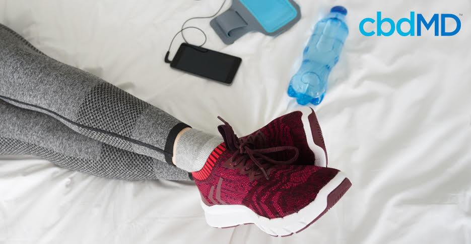 Athlete in gray leggings and red sneakers resting on bed with water bottle and phone nearby