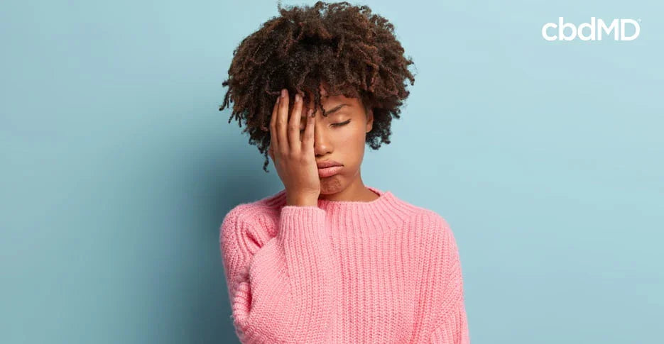 Woman in pink sweater looking stressed against blue background, hand on face, cbd wellness
