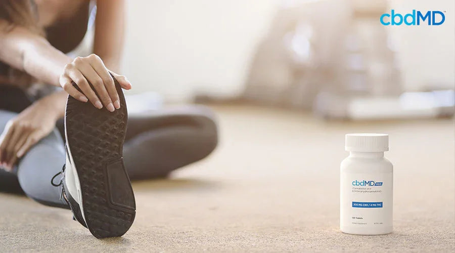 A woman stretching while sitting on the ground with max for pain next to her - cbdMD