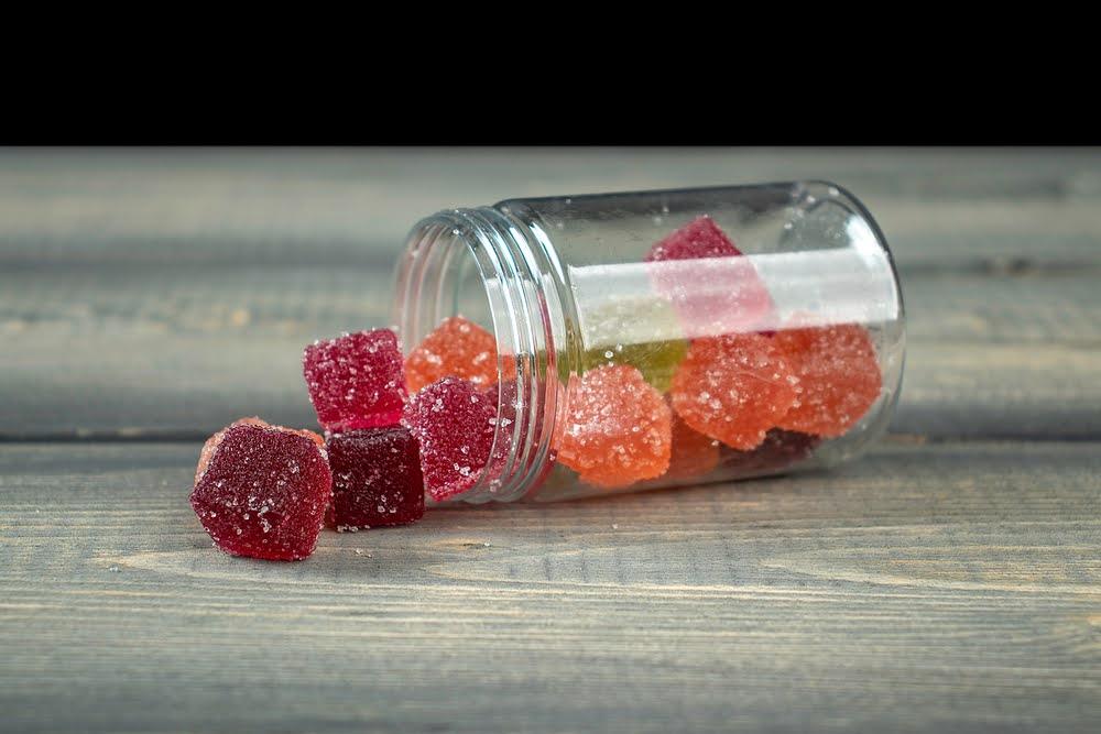 Assorted CBD gummies with sugar coating spilling from a clear jar onto a wooden surface
