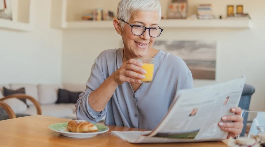 Smiling older woman with short gray hair reads newspaper and drinks orange juice at kitchen table