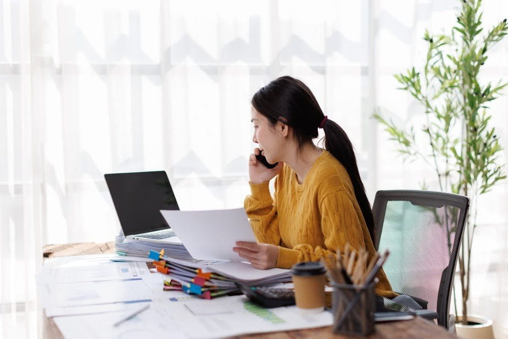 busy woman taking a call at her desk covered in papers and an open laptop