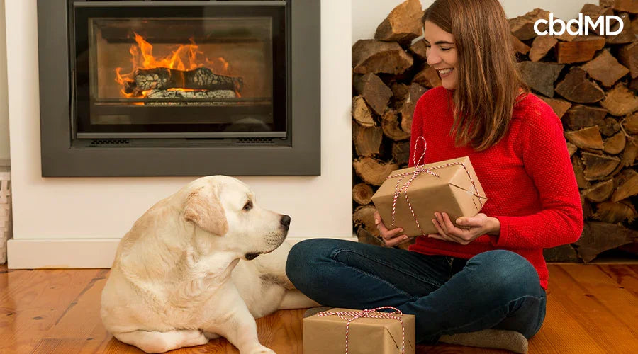 A woman holding a gift sitting down next to her dog laying down with another gift in front of them - cbdMD