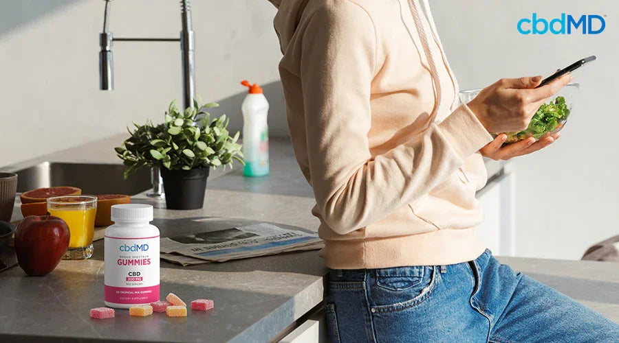 A woman in a kitchen holding a bowl of salad and her phone with broad spectrum gummies tropical mix on the counter - cbdMD