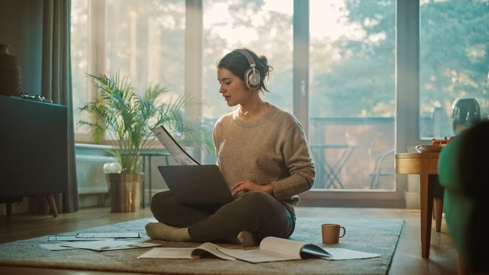 woman with headphones and a laptop in her lap sitting on the floor surrounded by papers