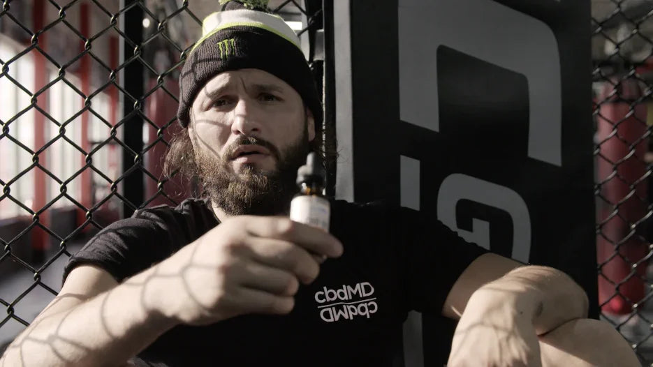 Bearded man in gym holding CBD oil dropper bottle, wearing black t-shirt and beanie