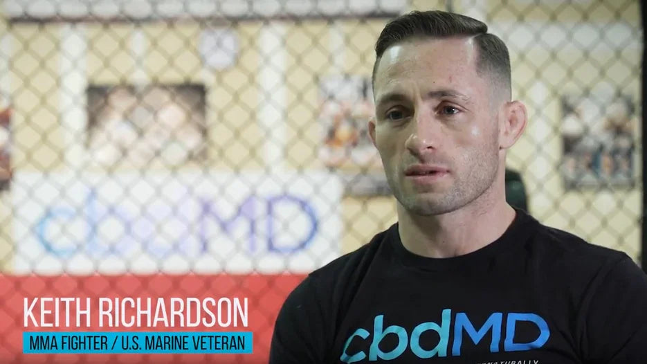 MMA fighter in black cbdMD shirt sitting in gym with chain-link fence background