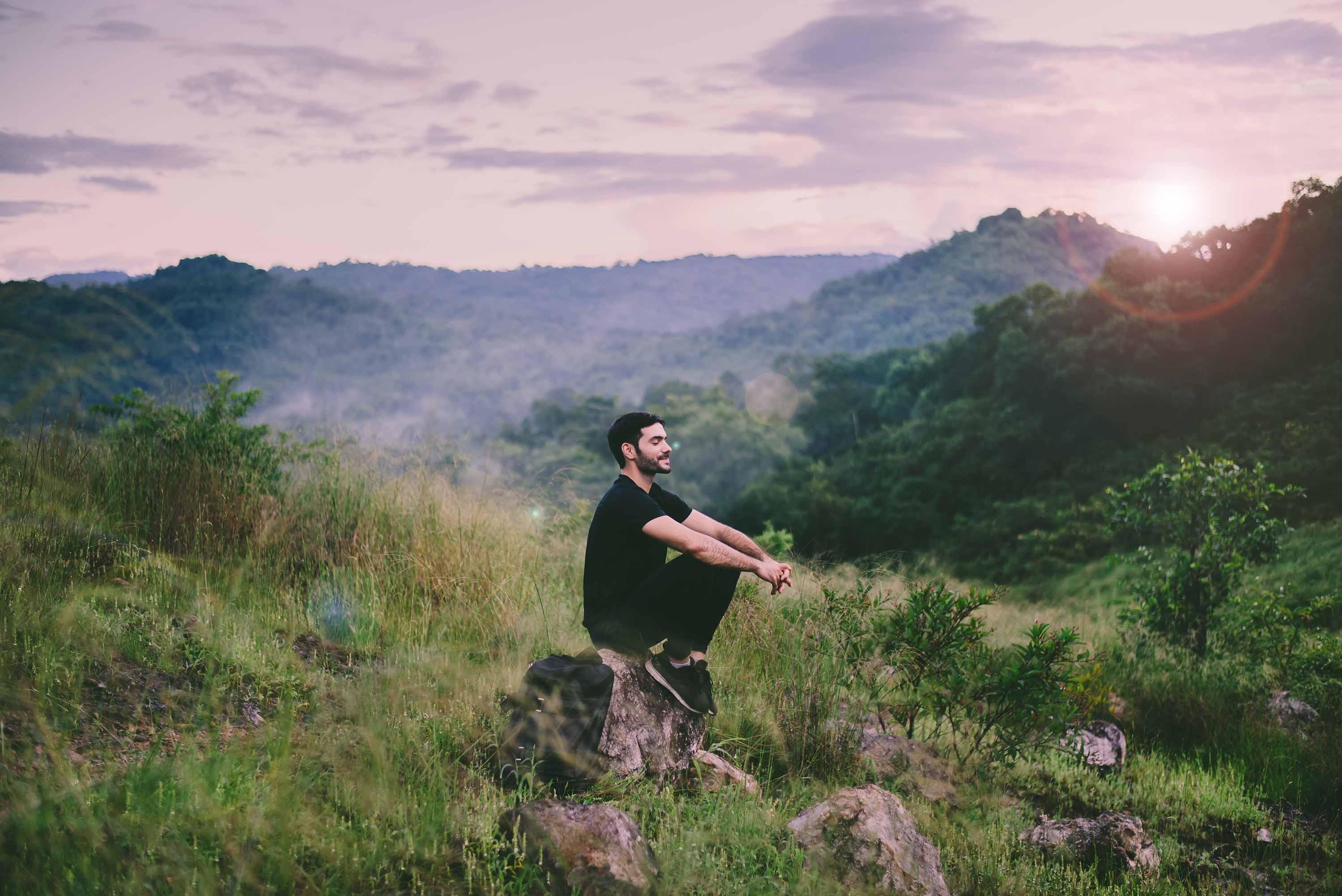 Man meditating on a grassy hillside at sunrise with peaceful mountain scenery
