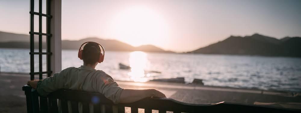 man wearing headphones and looking out from a bench over the water during sunrise