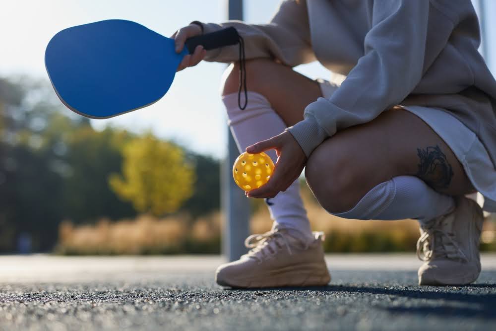 Person holding blue pickleball paddle and yellow pickleball on outdoor court