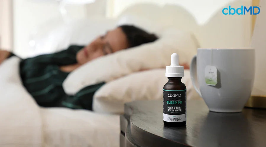 A woman sleeping in a bed next to a nightstand with full spectrum sleep pm 50 mg cbd 2 mg thc mint and a mug with tea on it - cbdMD