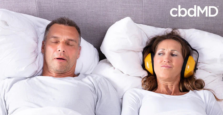 Woman in bed wearing ear protection looks annoyed at man snoring next to her, sleep issues concept