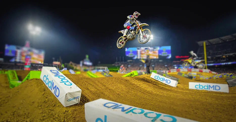 Motocross rider jumping dirt ramp at night supercross event with sponsor signs on track