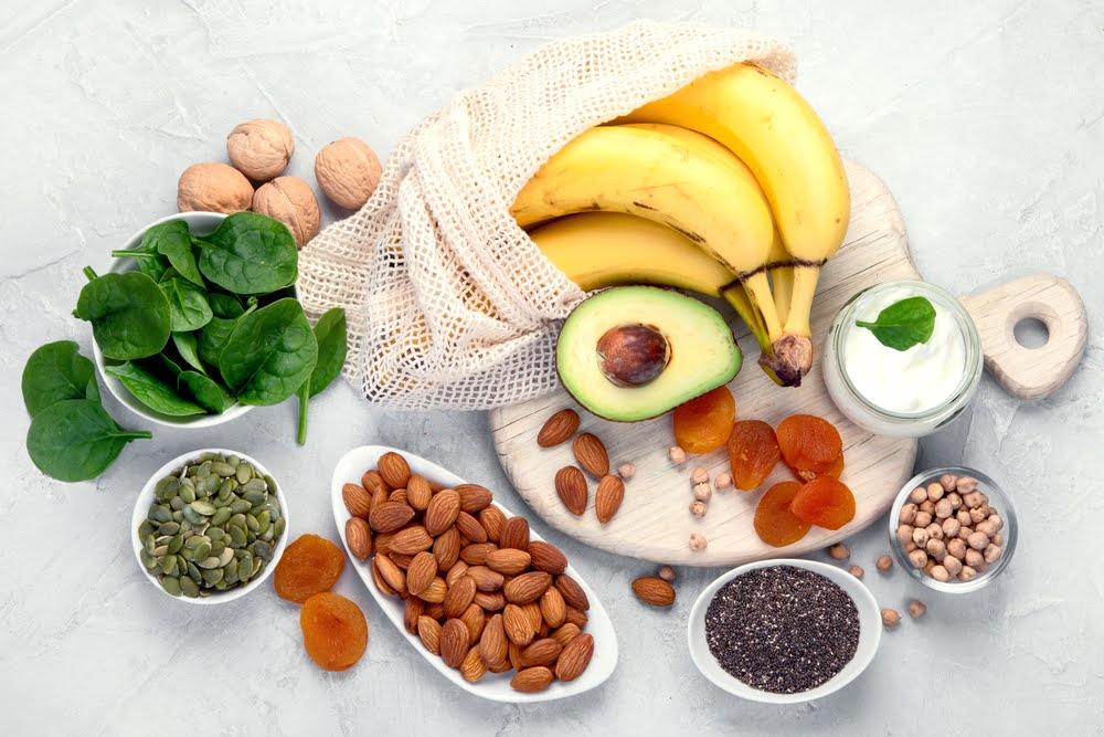 Healthy superfoods including spinach, bananas, avocado, nuts, almonds, chia seeds, yogurt, and dried apricots on a light background.