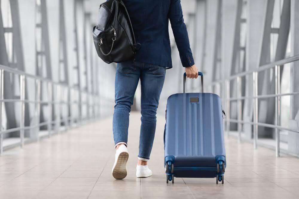 man walking away from the camera perspective in an airport concourse with a carryon rolling luggage