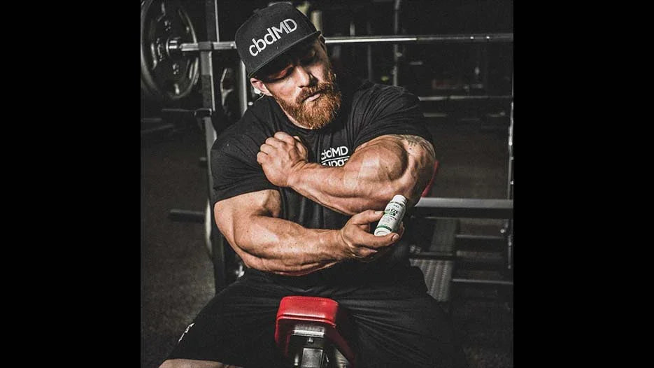 Muscular man in gym applying topical cream to arm, wearing cbdMD shirt and hat