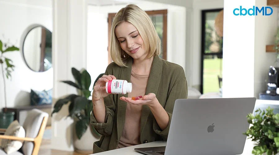 Woman at home pouring CBD gummies into hand near a laptop, relaxed and smiling