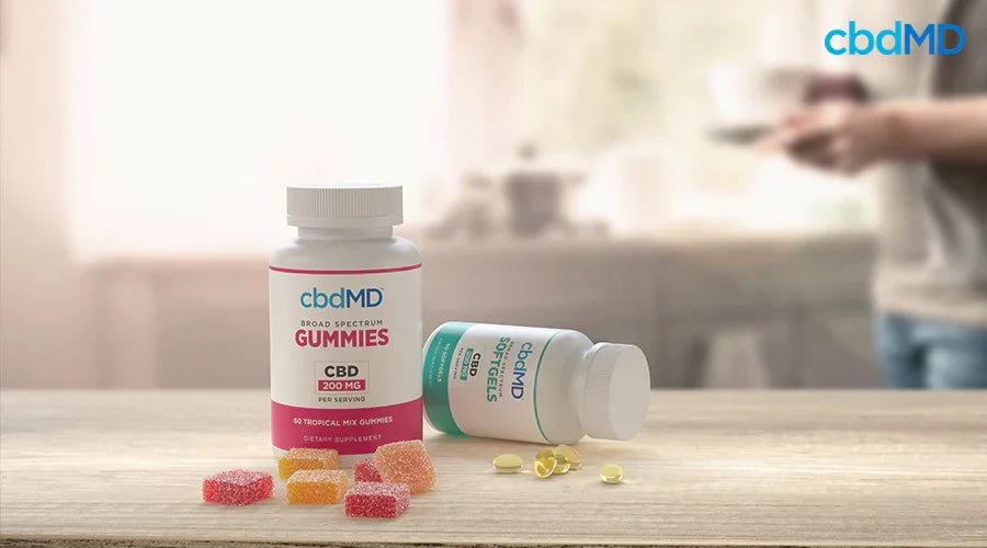 Broad spectrum gummies 200 mg per serving tropical mix and softgels on a kitchen counter with a woman in the background - cbdMD