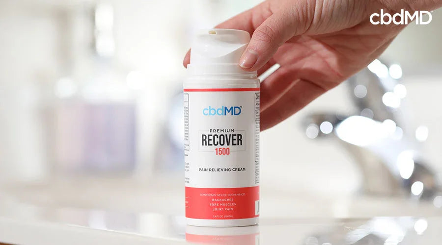 Hand holding a bottle of premium recover pain relieving cream in a bathroom setting.