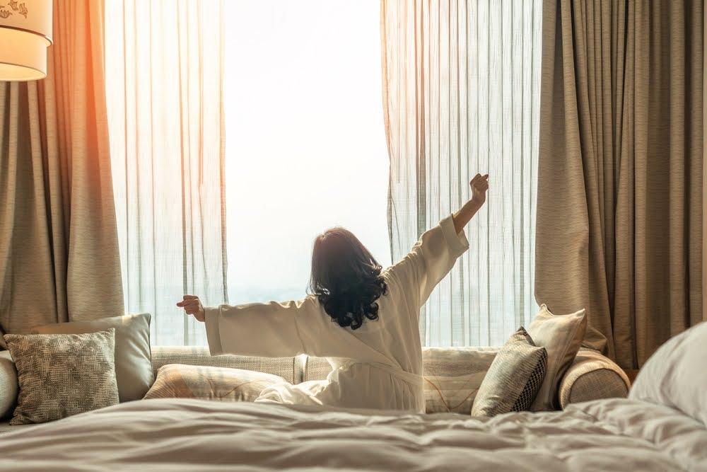 Woman stretching in bed by window with morning sunlight, wellness and relaxation concept